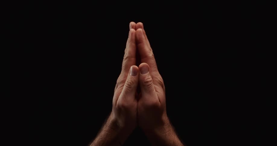 A man's hands slowly come together in a praying position against a stark black background