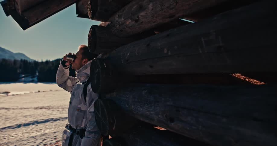 A man dressed in winter gear takes a break, drinking from a metal cup while leaning against a log cabin in a snowy, sunny landscape