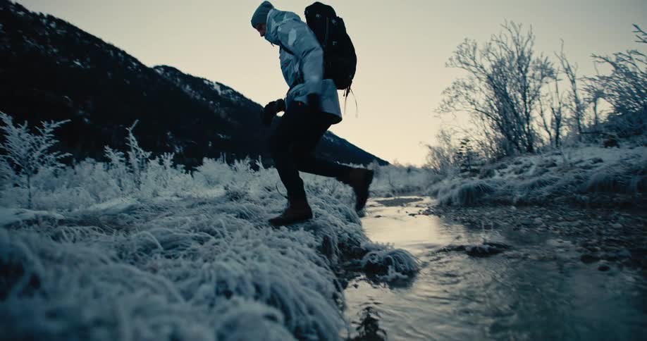 Slow motion shot of a hiker in winter gear carefully crossing a stream in a frosty, snow-covered landscape surrounded by mountains