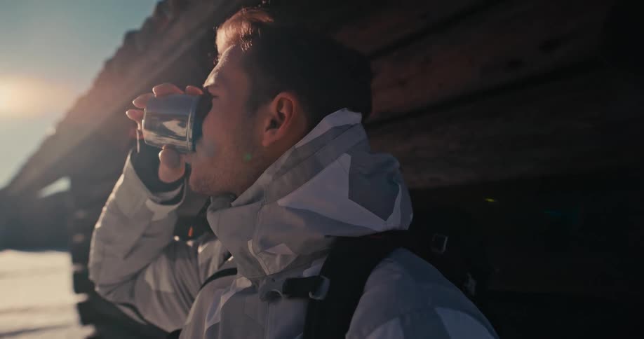 A male hiker enjoys a hot drink from a thermos while resting against a log cabin in a sunny, snowy mountain landscape
