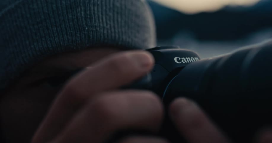 A male photographer in a beanie takes a picture with his camera, then lowers it to thoughtfully gaze at the scenic landscape