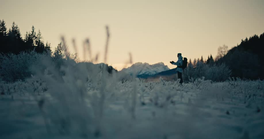 A photographer with a backpack stands in a frosty, snow-covered field to capture the beautiful mountain landscape at dawn
