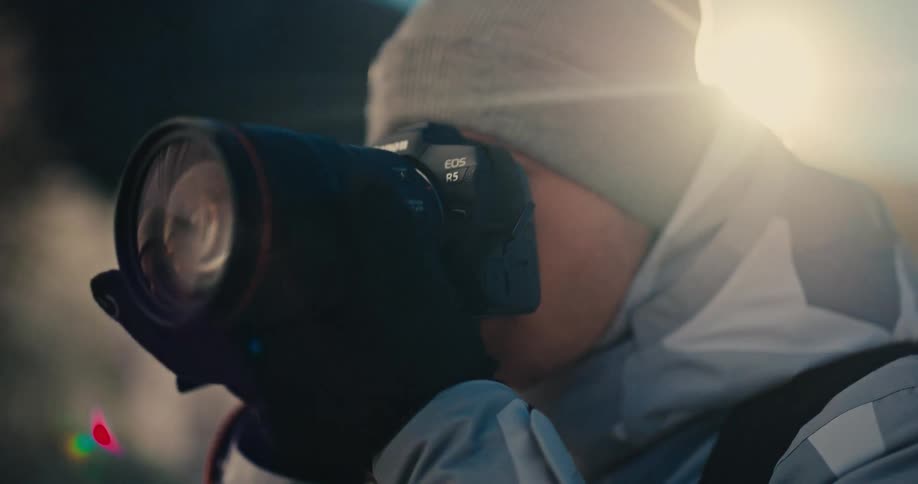 A professional photographer bundled in winter gear captures a moment with his camera in slow motion, backlit by the golden sun
