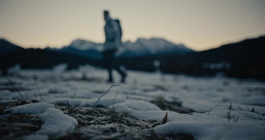 A close-up on frosty ground as a lone hiker walks through a snowy mountain landscape at dusk