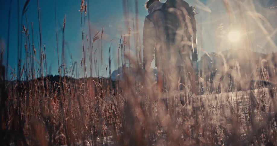 A lone hiker with a backpack walks in slow motion through a sun-drenched, snowy meadow with tall grass towards distant mountains