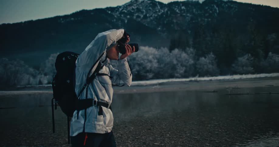 An adventurous photographer in a winter jacket captures the moody, misty mountain landscape by a river at dawn