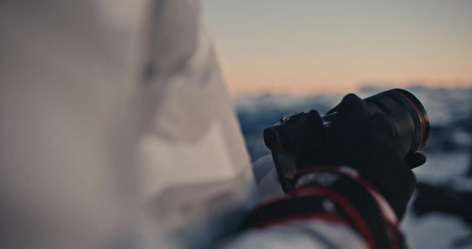 An over-the-shoulder shot of a photographer in a white jacket and gloves capturing the sunrise over a snowy mountain range