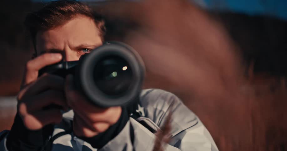 A cinematic close-up shot shows a photographer pointing his camera directly at the viewer as the focus pulls from the lens to his face