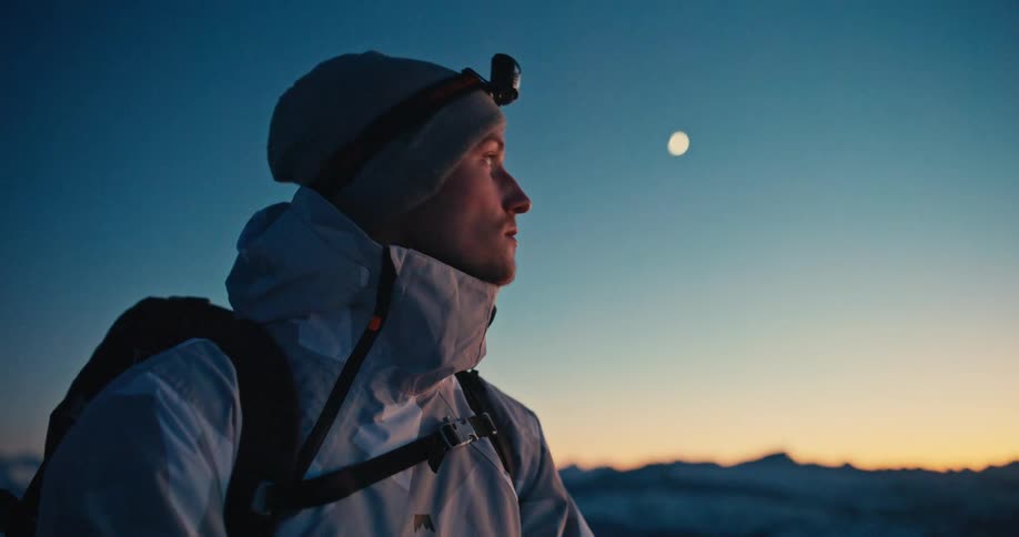 A male mountaineer with a headlamp looks out at the moon and the serene twilight sky over a snowy mountain landscape