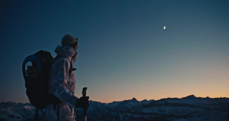 A hiker in full winter gear stops on a snowy mountain ridge at dusk, taking in the epic view of the mountain range and the crescent moon above