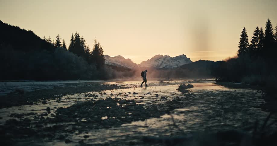 A silhouetted adventurer walks through a misty, shallow river in a vast mountain landscape at sunrise