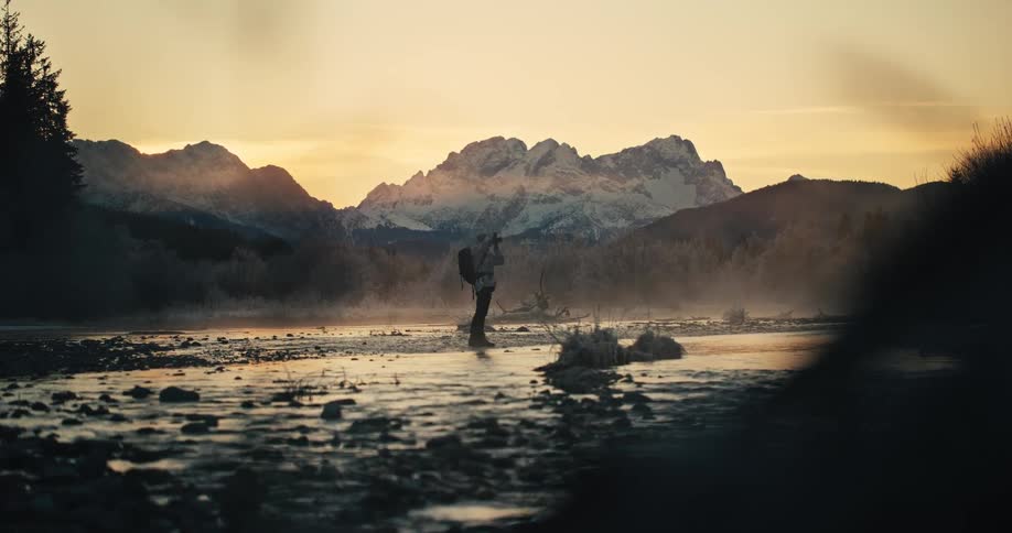 A photographer with a backpack stands in a misty river taking pictures of the stunning snow-capped mountains at sunrise