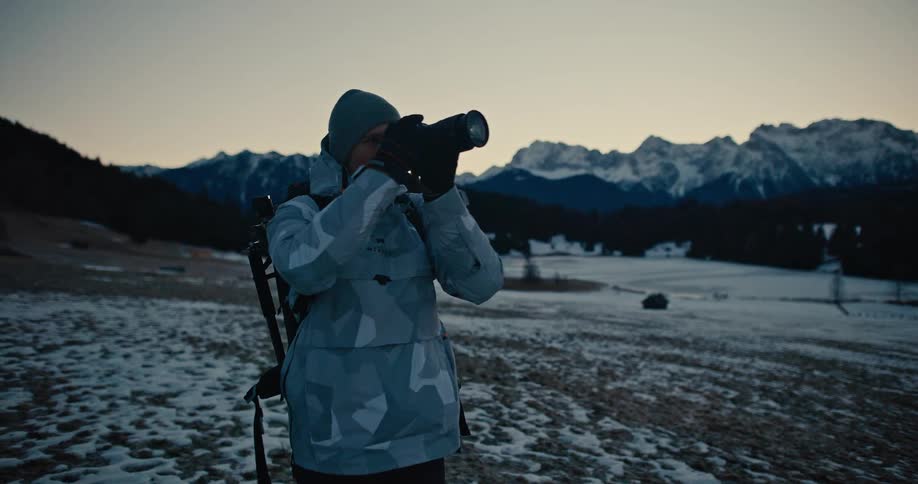 A male photographer in a snowy alpine landscape raises his camera to capture the majestic mountain views at dusk