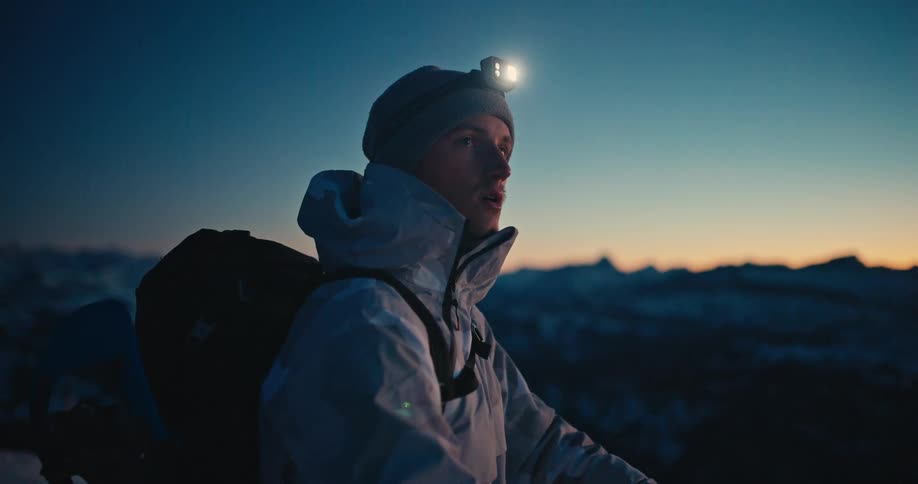 A mountaineer with a headlamp gazes at the snowy mountain peaks as twilight settles over the landscape