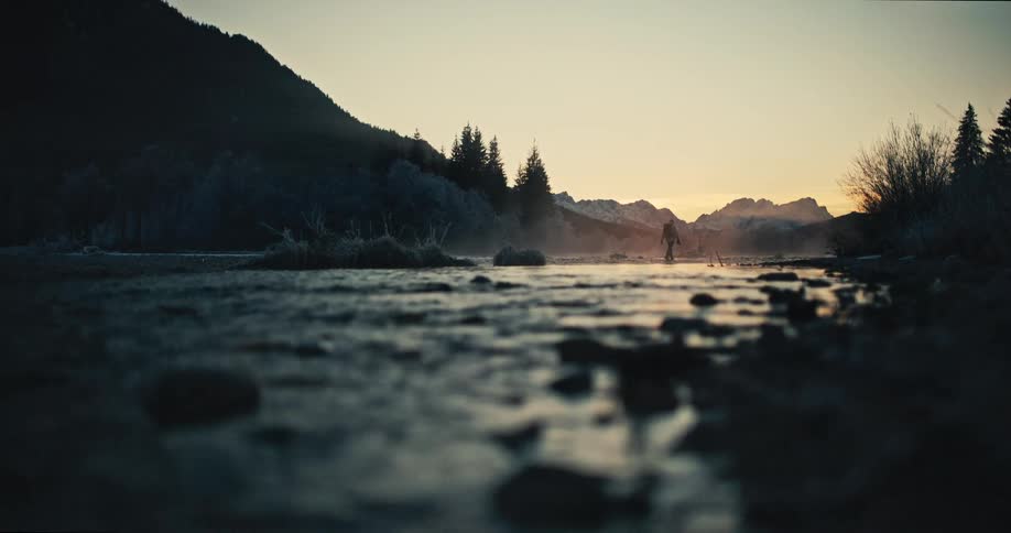 A cinematic low angle shot captures a lone hiker standing silhouetted in a misty river surrounded by mountains at dawn