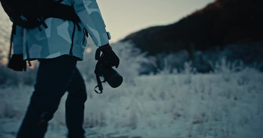 A photographer hikes through a frosty, snow-covered landscape at dawn in slow motion