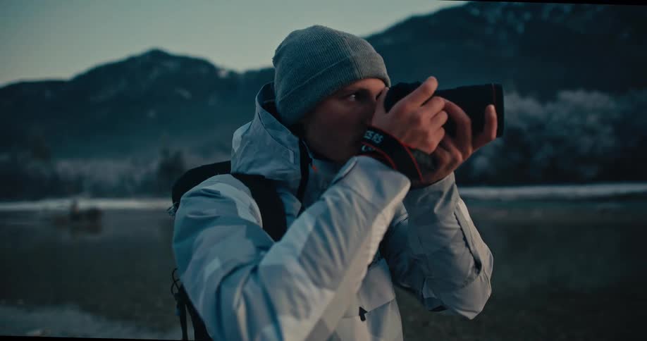 A male photographer with a backpack and winter gear takes a photo of a frosty mountain lake before admiring the view in slow motion