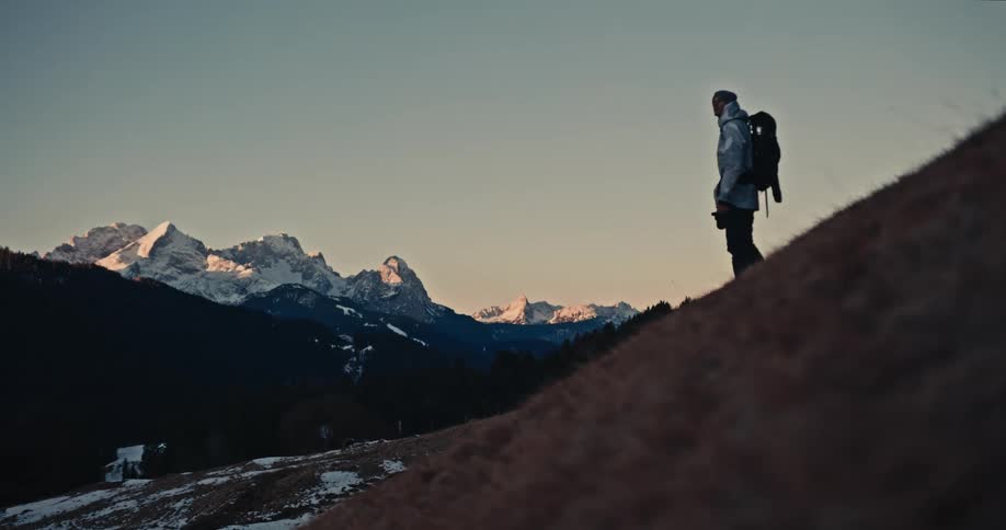 A lone photographer hikes up a grassy, snowy hill at sunrise to capture the magnificent mountain range view