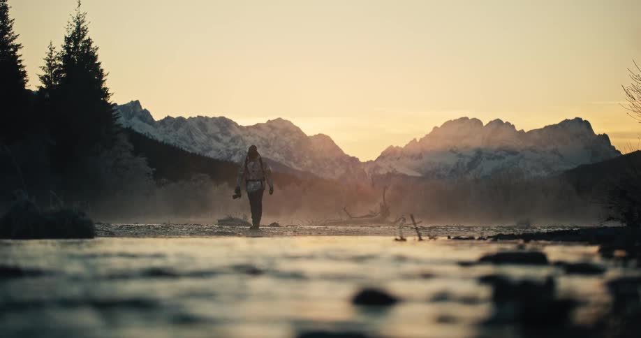 An adventurer with a camera walks through a misty river in a majestic mountain valley during a golden sunrise