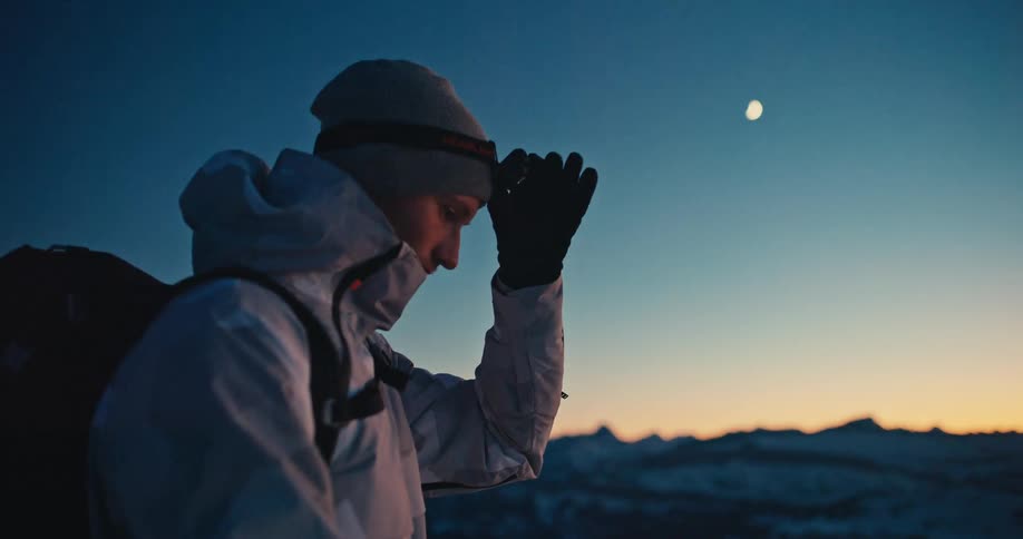 A male mountaineer turns on his headlamp as he gazes at the crescent moon over a snowy mountain range at twilight