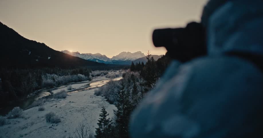A photographer takes a picture of the sun rising over a majestic, snowy mountain range and frosty river valley
