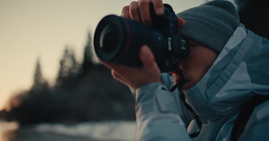 A focused photographer in a winter jacket and beanie raises his camera to take a picture in a cold, misty landscape during twilight