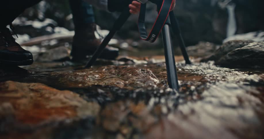 A nature photographer carefully places a tripod into the clear water of a rocky stream in super slow motion