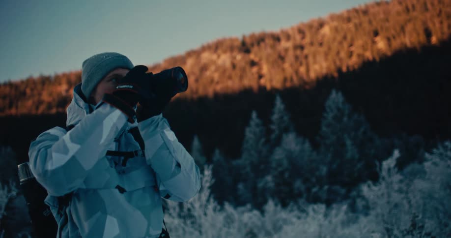 A male photographer in a camouflage winter jacket stands in a snowy landscape and takes photos of the mountains during golden hour