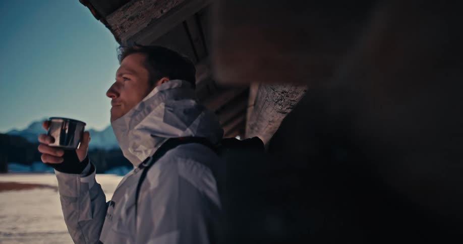 Slow motion shot of a man in a white winter jacket drinking from a metal mug, enjoying the sunny, snowy mountain landscape from a rustic cabin