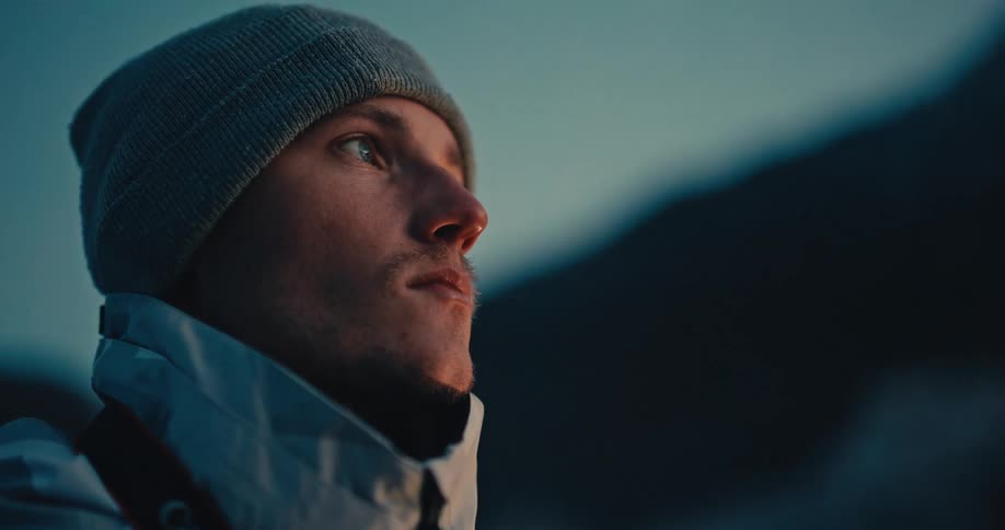 A close-up of a pensive young man in a beanie and jacket looking into the distance, his face lit by a warm glow at dusk