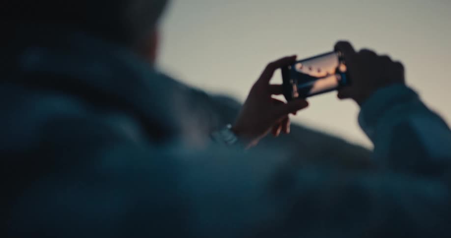An over-the-shoulder shot shows a person taking a scenic photo of snowy mountains and a crescent moon with their smartphone at dusk