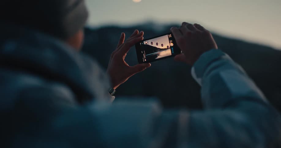 Over the shoulder shot of a person taking a scenic photo of a mountain lake with their smartphone at dusk