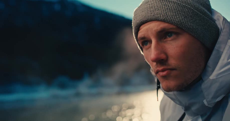 A focused photographer in winter clothes adjusts his camera on a tripod by a steaming lake at sunrise