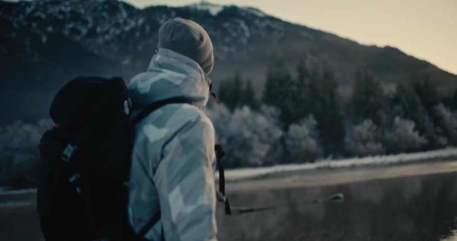 A hiker in a winter jacket walks in slow motion along a misty, frozen lake, surrounded by snowy mountains and frosty trees