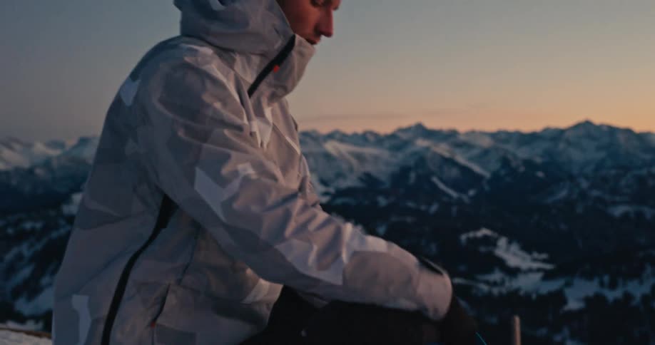 A solitary mountaineer unpacks his camera from his backpack on a snowy mountain summit at sunset