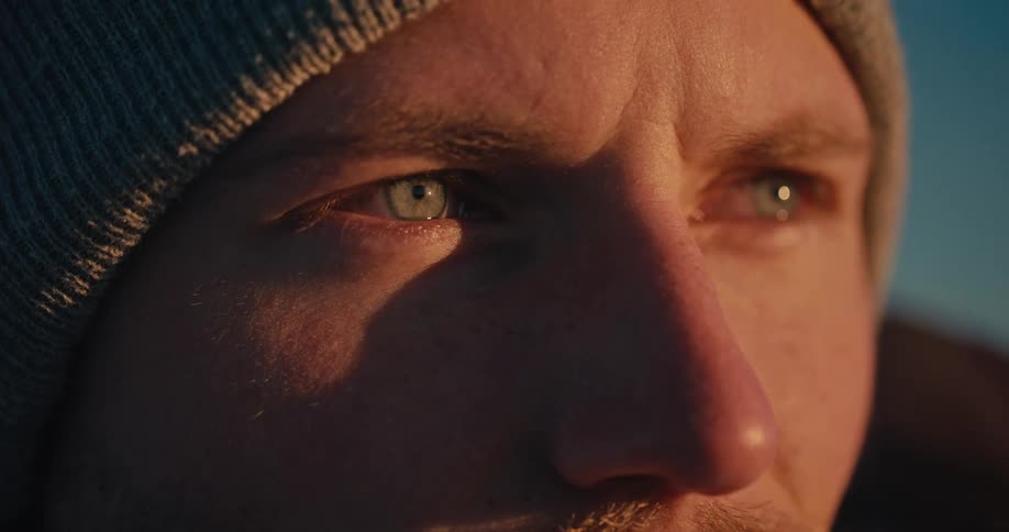 An extreme close-up reveals the focused, blue eyes of a man in a beanie, illuminated by the warm glow of a sunrise