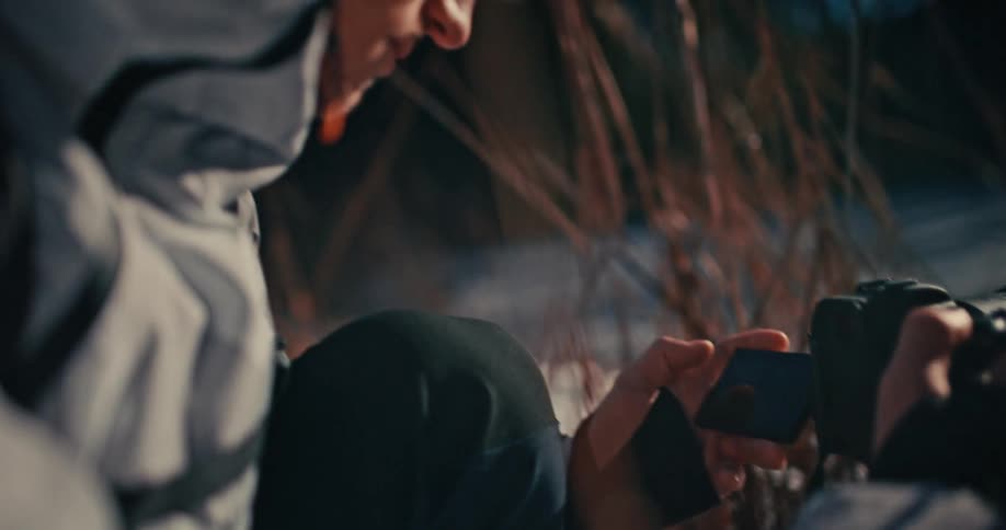 Two filmmakers collaborate on a low-angle shot amidst sun-drenched, snowy reeds in a close-up, over-the-shoulder view