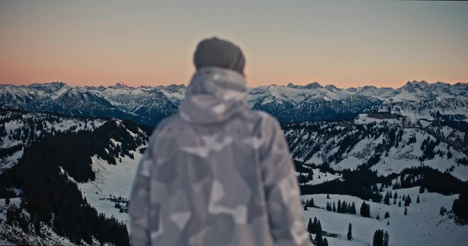A person in winter gear gazes out at a panoramic vista of snow-covered mountains during a serene sunset