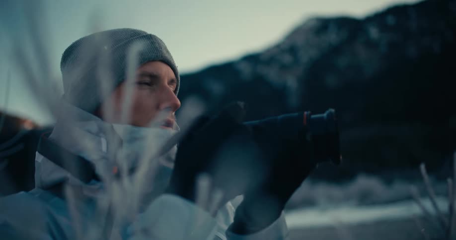 A focused photographer in winter gear captures a frosty mountain landscape at dusk in slow motion