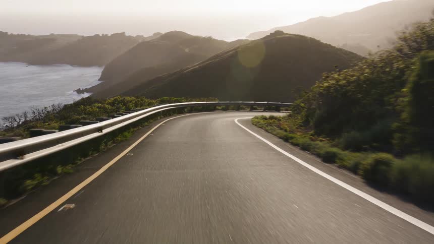 A point-of-view shot of a car driving on a scenic, winding mountain road along the coast at sunset
