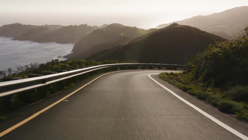 A first-person point of view driving on a winding road through lush green hills along the coast during a beautiful golden sunset
