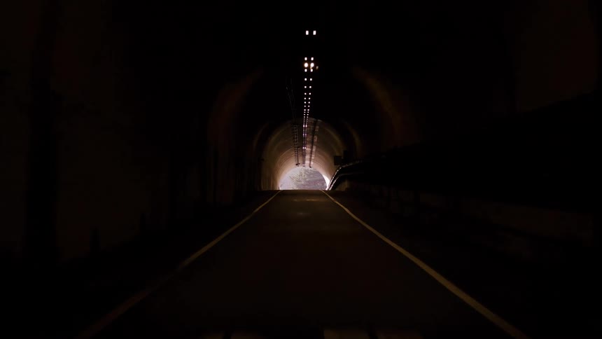 A point-of-view shot driving slowly through a dark tunnel and emerging into the bright sunshine on a winding country road