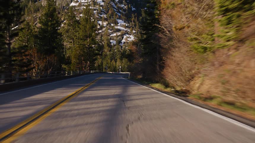 Point-of-view driving shot along a winding mountain road with snowy peaks and evergreen forests on a sunny day