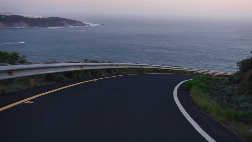 A scenic point-of-view drive along a winding coastal road overlooking the ocean and a distant city skyline at dusk
