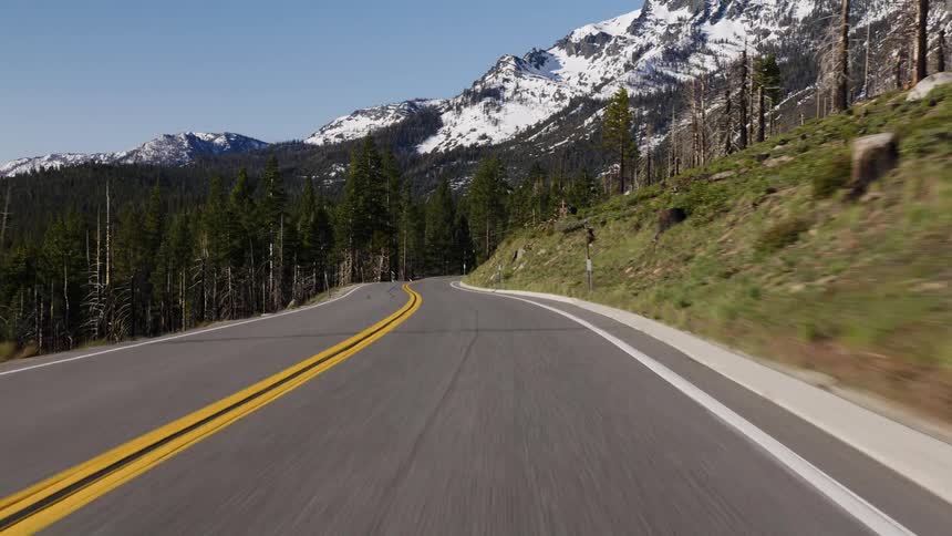 Point-of-view shot of driving along a scenic winding mountain road with snow-capped peaks and a pine forest on a sunny day