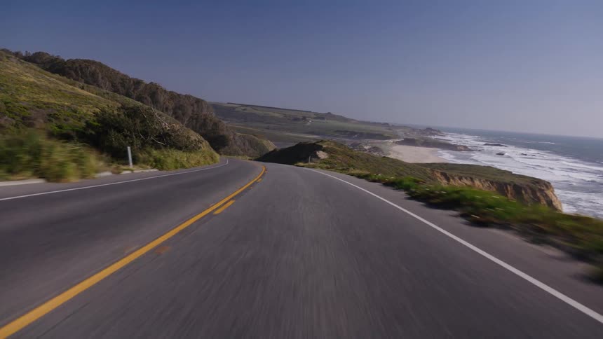 A scenic point-of-view drive along a winding coastal highway with the ocean on the right and green hills on the left