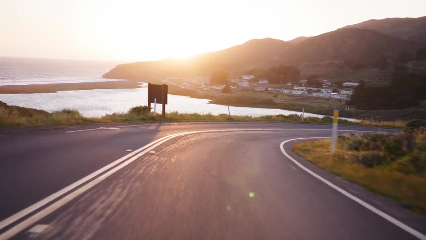A point-of-view shot of driving along a winding coastal road through rolling hills at sunset