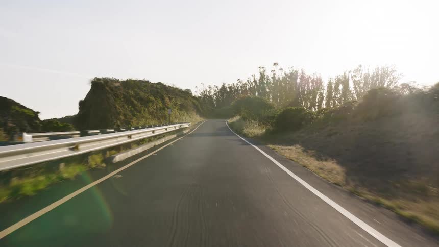 A slow, forward-moving point-of-view shot driving along a winding asphalt road through a grassy, hilly landscape on a sunny day