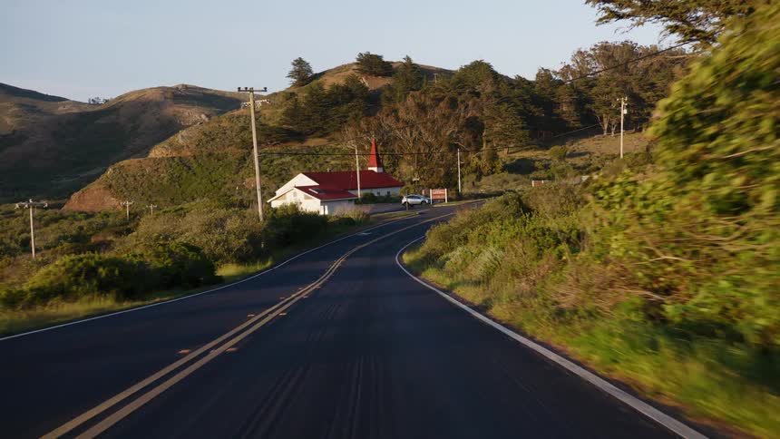 A serene, smooth forward-moving shot down a winding country road leading to a quaint church nestled in the rolling green hills at golden hour
