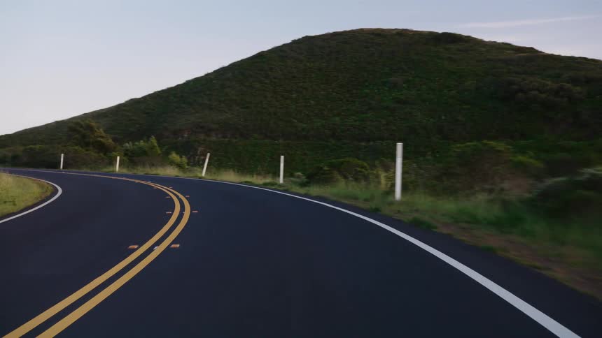 A smooth point-of-view shot follows a curving paved road through lush green hills at twilight